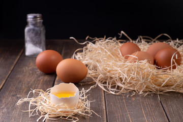 An open egg with the white and yolk on the hay, in the background in a nest of hay are whole fresh eggs, salt. Wooden background. Copy space. Space for text.