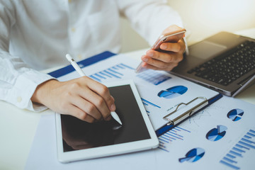 Businessman holding smartphone and using a pen to write on the tablet while he is working with laptop to analze business graph chart on the desk in the office.