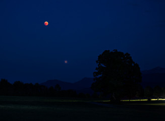Blood moon eclipse during summer 2018 with planet Mars above fields in the night sky, stars and...