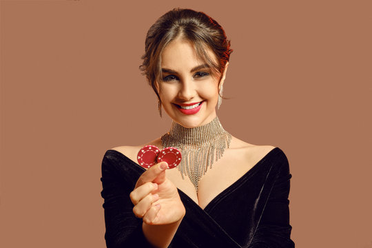Brunette model in black dress and shiny jewelry. She smiling, showing two red chips, posing on brown background. Poker, casino. Close-up