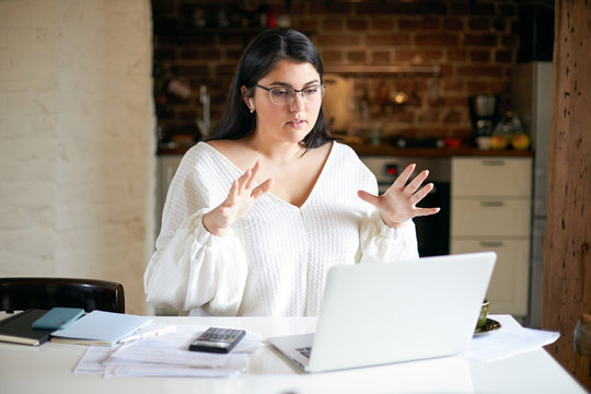 Serious Young Plus Size Businesswoman In Wireless Earbuds Sitting In Front Of Open Laptop, Gesturing Emotionally, Discussing Business Plans And Development Strategy With Team Via Video Conference Chat