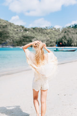 A thin young girl posing along in a white cape the sea. The blonde in a knitted lace shorts and beige t-shirt. White sand beach, clear turquoise & azure water in Thailand. Chic hippie fashion.