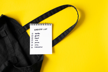 Shopping or grocery list in paper spiral notepad on empty cotton eco black bag on yellow background...