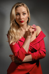 Blonde woman with bare shoulder, in red dress and black earrings. She showing two chips, posing on gray background. Poker, casino. Close-up