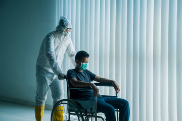 Doctor with hazardous materials suit pushing a patient infected with Coronavirus in a wheel chair