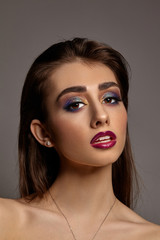 Naked woman in necklace and earrings posing on gray background. Luxury makeup, perfect skin. Colorful eyeshadow, long eyelashes, purple lips. Close up