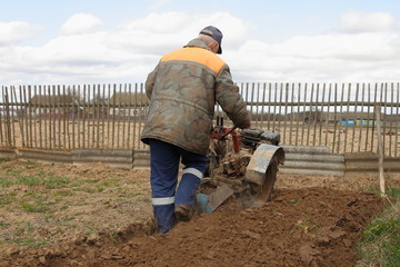 The farmer in the warm work clothes plows tillers of the ground  on old wooden fence background at spring day