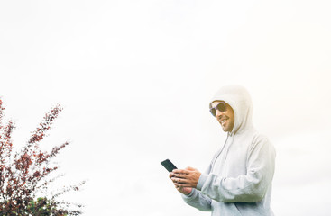 Young man in gray sweatshirt and blue sunglasses holding cell phone in his hands and smiling