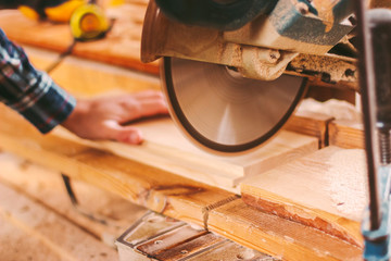 Closeup of professional joiner working with electric circular saw at woodworking workshop. Skilled man cabinet maker using circular saw for cutting wooden board at sawmill. Wood production factory