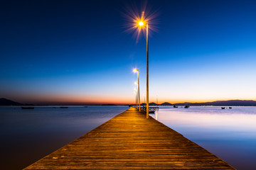 wooden pier in calm sea at sunrise, blue hour, fishing boats and land on the horizon - 343577014