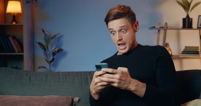 Young handsome man eading shocking news while using his smartphone . Cheerful guy in 30s making amazed face and big eyes while looking at phone screen and sitting on sofa at home.