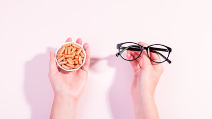 Woman holding eyeglasses in one hand and vitamins for eye health in other. How to maintain good eyesight and keep eyes healthy concept. Top view, pastel background