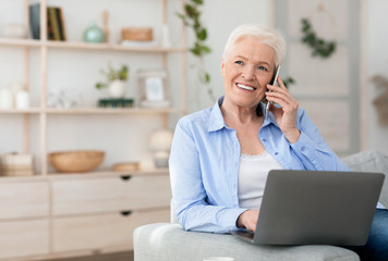 Modern senior woman using laptop and talking on cellphone at home