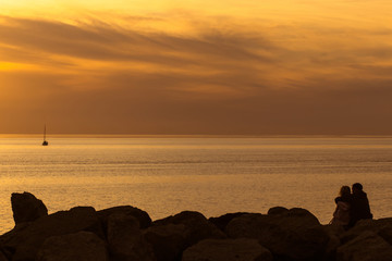 Silhouettes of loving couple are sitting on the stones closeup at sunset time near the sea.