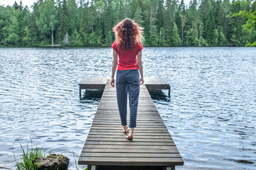 Fototapeta premium Young girl walking on the pier, back to the camera. A view of the beautiful lake. Summer mood.