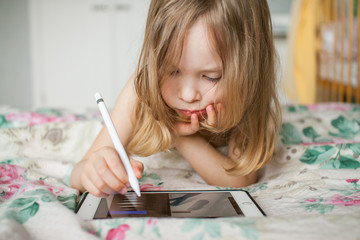 A little girl painstakingly and enthusiastically draws with a stylus on a tablet at home. Modern...