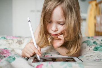 A little girl painstakingly and enthusiastically draws with a stylus on a tablet at home. Modern...