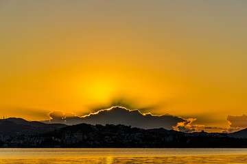scenery with sun rising behind the clouds over an urban landscape, with golden sea in the foreground - 343583451