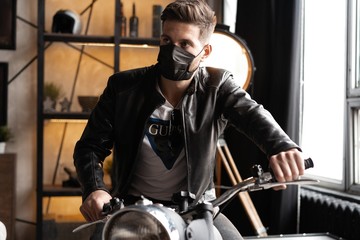 Fototapeta premium Handsome brutal male biker in black mask in leather jacket sitting on motorcycle looking forward.