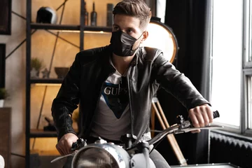 Foto op Plexiglas Handsome brutal male biker in black mask in leather jacket sitting on motorcycle looking forward. © opolja