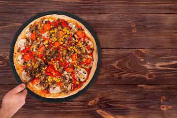 Man cut the pizza with a kitchen knife. Pizza with mushrooms, corn, cherry tomatos, courgettes and bell peppers or Veggie Vegetarian pizza on the slate plate on wooden background, top view