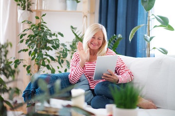 Attractive senior woman with tablet sitting indoors on sofa, video call concept.