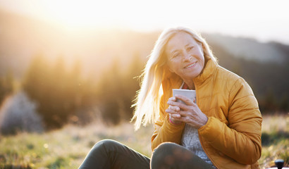 Attractive senior woman sitting outdoors in nature at sunset, relaxing with coffee.