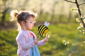 Small toddler girl standing outdoors in orchard in spring, holding paper bee.