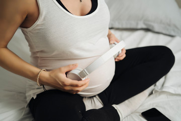 Cropped image of beautiful pregnant woman playing music with headphones on her belly to relax baby sitting on bed in her room