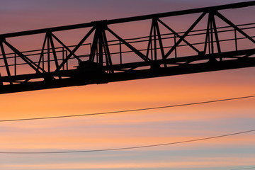 Fototapeta na wymiar The shape or silhouette of a tower crane against the sky and the setting sun. Bridge crane at the factory in the evening. Beautiful cloud landscape in the form of horizontal lines of yellow and orange