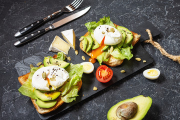 Healthy sandwich - poached eggs and avocado on toast with tomatoes on black stone background.