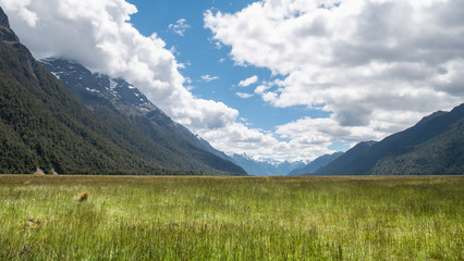 Beautiful picturesque valley surrounded by mountains with green fields in foreground and blue sky with some clouds in background. Photo taken in Eglinton Valley, Fiordland National Park, New Zealand