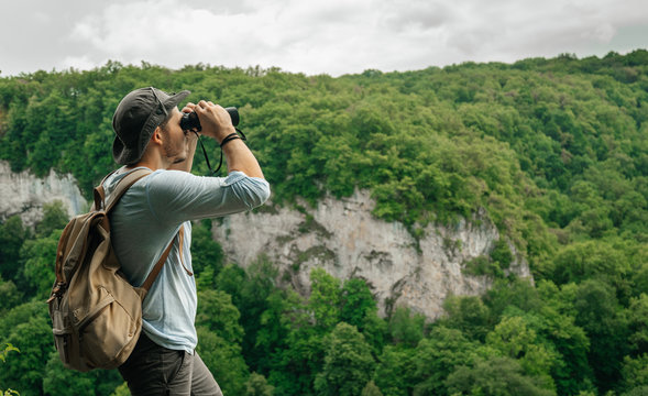 A man uses binoculars while standing at a height with a backpack on his back inspecting the forest. Environmental studies and forest fire protection.