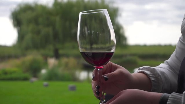 A woman in white garment with manicured hands holding a glass of red wine at outdoor restaurant. A business woman having a drink and a talk in a summer garden. Bright green grass and a lake in a park.