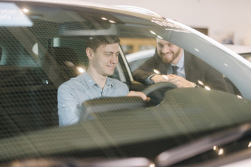 Happy young man behind the wheel of new car in auto dealership. Professional car salesman wearing business suit is telling about new car model. Concept of choosing and buying new car at showroom.