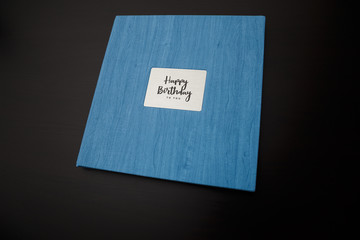 Photobook, photoalbum in blue leather cover on black table background with metallic shield and inscription Happy Birthday to you. Top view, copy space.