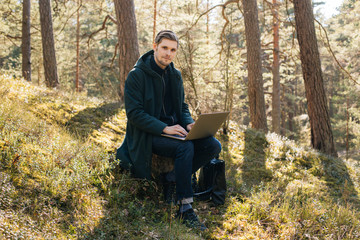 Obraz premium Man using a laptop work in forest. Young man sitting on an old tree strain work remotely in nature. Work everywhere traveler blogger outdoors. Businessman sitting in forest Remote working concept 