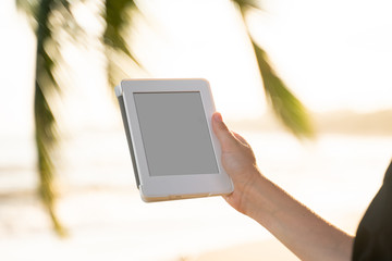 Woman holding e-reader outdoors by the sea with a palm trees on background. Relaxing and enjoying with a reading a favorite books outdoor in a travel.