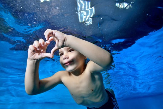 A beautiful little boy swims underwater in a children's pool with his eyes open and makes a heart shape out of his fingers. Closeup. Concept. Digital photo. Horizontal view.