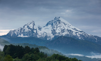 Obraz premium Snow covered mountain peaks of Watzmann at Berchtesgaden with clouds and morning fog, green forest in foreground, Bavaria.