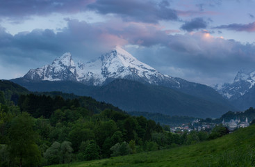 Snow covered mountain peaks of Watzmann and city Berchtesgaden with clouds during sunset, green forest and fields in foreground, Bavaria.