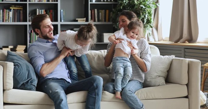 Joyful young family spending time together having fun tickling relaxing on sofa. Happy adult parents and two small cute children laughing playing, enjoying lifestyle activity in living room together.