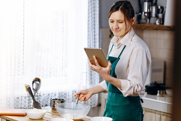 Fototapeta premium Woman looking to laptop and reading recipe for cooking dishes. Cooking at home concept, lifestyle. Preparing the dough, baking in the kitchen.