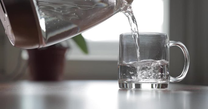 The glass transparent glass is on a table, water pour from a teapot in a glass, flowers are on background