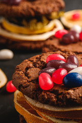 Homemade oat chocolate cookies sandwich with dried citrus fruits and juicy jelly beans on textured dark black background, angle view macro