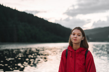 Pretty girl in a red raincoat stands on the background of a lake in the mountains and looks in camera with a serious face. Attractive hiker girl on an evening walk on background of mountains and lake