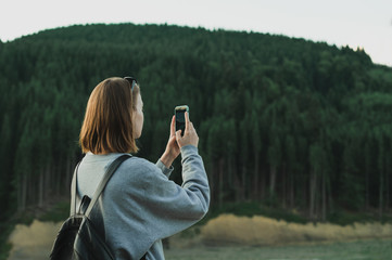 Pretty girl sitting on the apartment terrace on the background of mountain landscape and lake, using a smartphone with a serious face. Girl tourist uses internet on smartphone on balcony in mountain