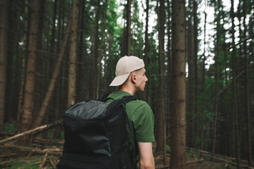 Naklejka na ściany i meble The back of a young man in casual clothes and with a backpack walks through the pristine coniferous forest and looks away. Guy tourist walks through the forest.