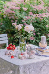 Pink peonies in a glass vase and books in lilac covers, sweets, marshmallows and ripe strawberries with a bowl on a white table in the garden