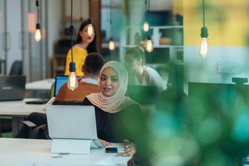 African muslim girl wearing hijab working on a laptop in a modern startup company. While being gossiped by her european coworkers.